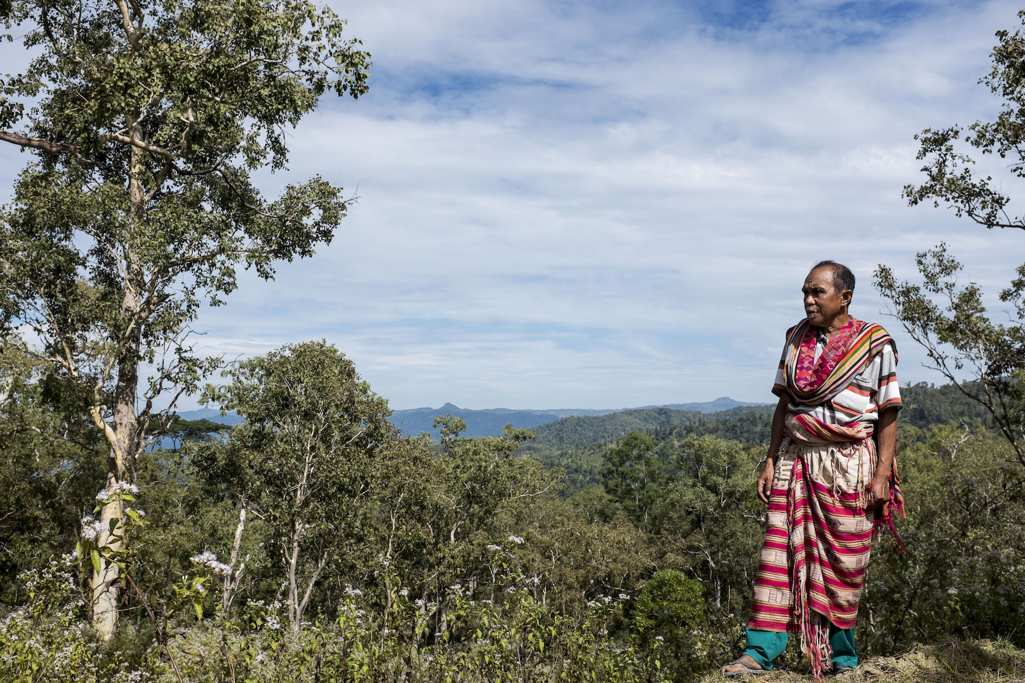 Isak Fobia stands on a hillside in West Timor, Indonesia, surrounded by forest and distant mountains.