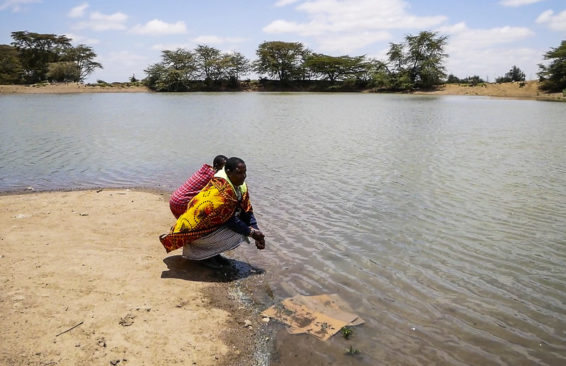 Two farmers crouch on the shoreline of a dam