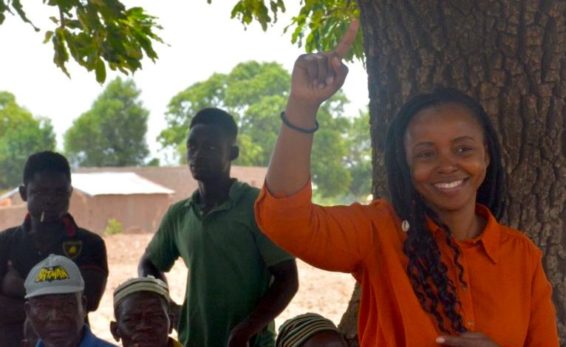 Woman stands beside tree, speaking with farmers
