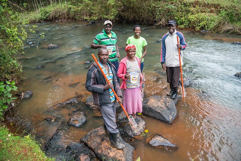 A group of five people stand on rocks in a river, some of them holding sticks with plastic water bottles attached to take samples