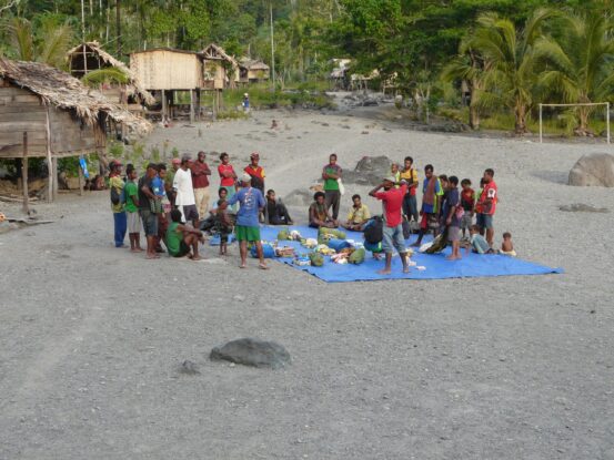 Man in traditional attire with a spear stands near a fire where bananas are roasting, while women and children sit nearby outside a stilt house on the Ghost Mountain Trail in Papua New Guinea.