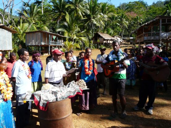 Group of villagers, some wearing flower garlands, singing and playing guitars outdoors in a rural village with traditional houses and palm trees along the Ghost Mountain Trail in Papua New Guinea.