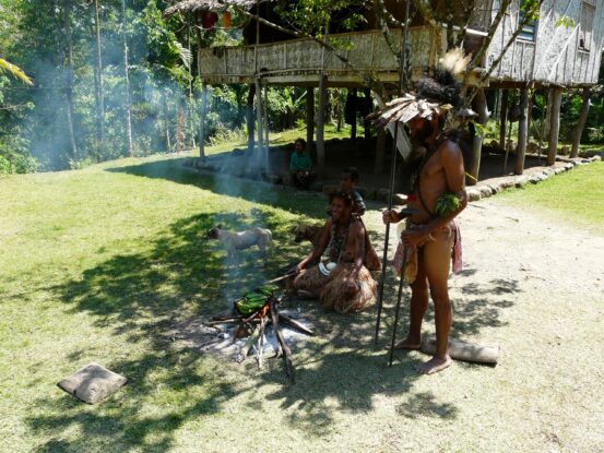 Man in traditional attire with a spear stands near a fire where bananas are roasting, while women and children sit nearby outside a stilt house on the Ghost Mountain Trail in Papua New Guinea.