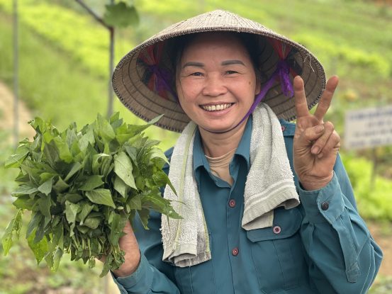 A smiling Vietnamese woman family farmer wearing a conical hat holds freshly picked greens on her agroecological farm in Hòa Bình, demonstrating how family farmers and agroecology support sustainable livelihoods.