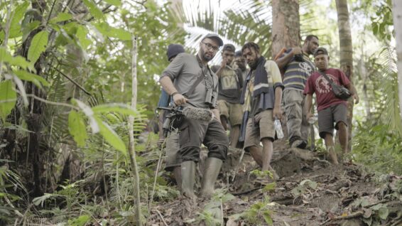 Gabriel Leite stands in the foreground wearing glasses, a cap and rubber boots while holding a directional microphone with a furry windscreen as a group of local men follow him down a muddy jungle path.