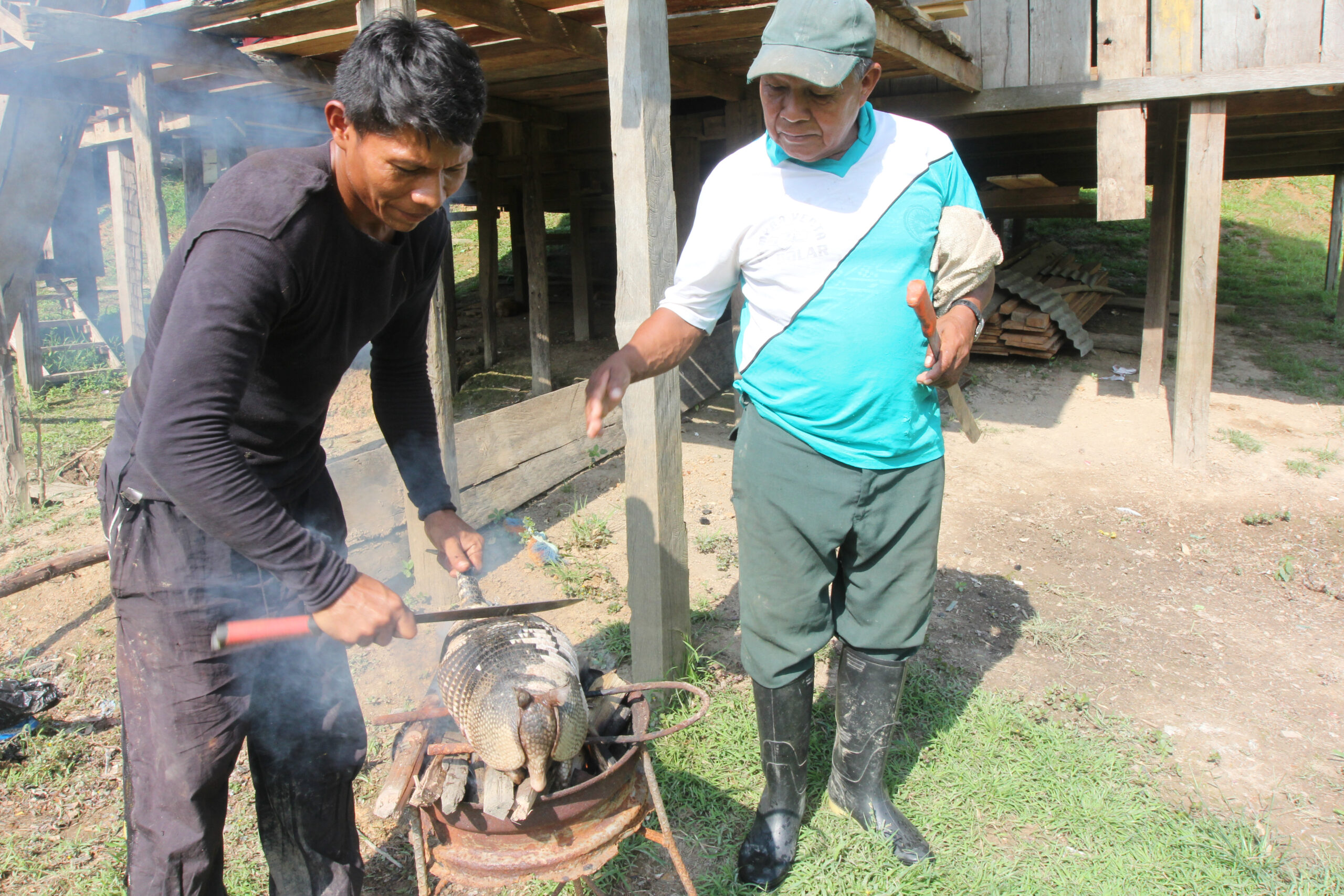A hunter prepares a trapped armadillo over an outdoor wood fire while a neighbour watches near a wooden stilt house.