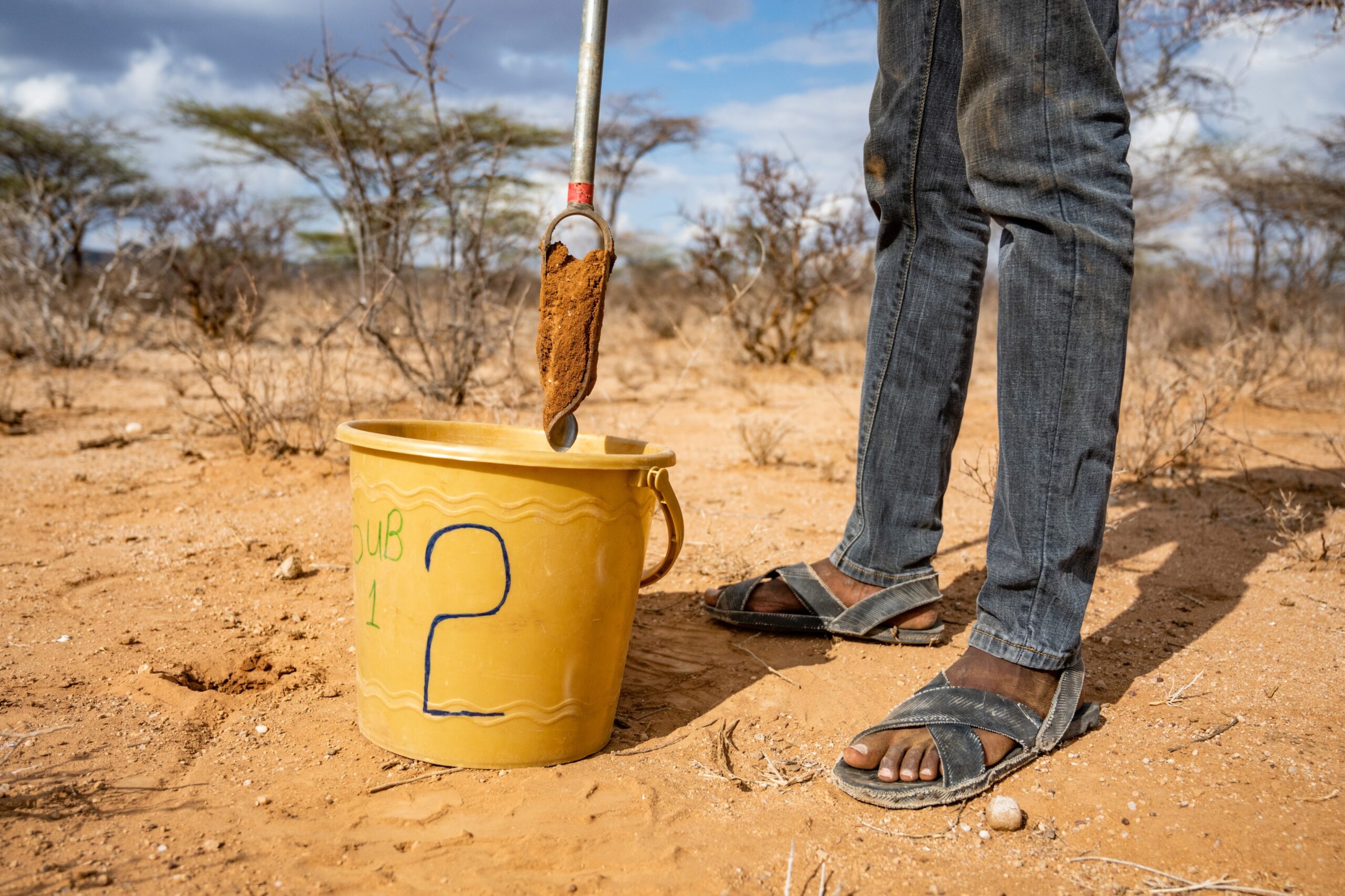 A field researcher lifts an orange soil sample over a yellow collection bucket in a dry area.