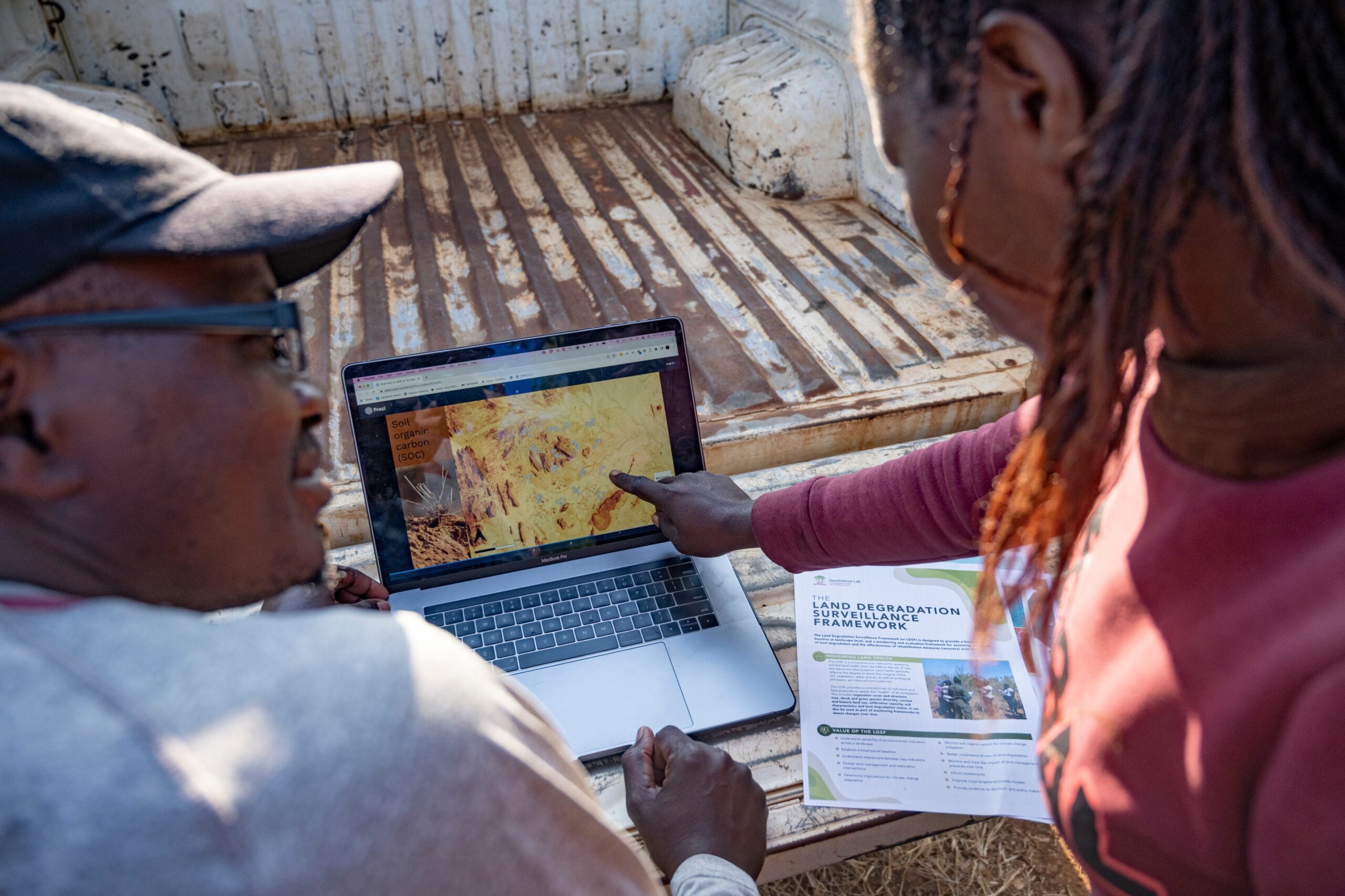 Two people examine soil information displayed on a laptop beside a land-monitoring guide.