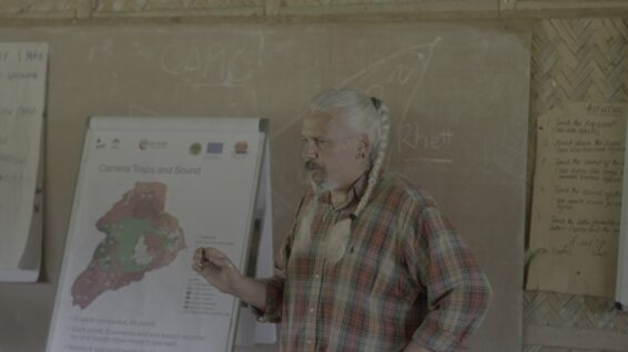 Ecologist Rhett Harrison, with long grey hair in braids and wearing a plaid shirt, gestures while standing next to a presentation board displaying a map titled "Camera Traps and Sound" inside a classroom.