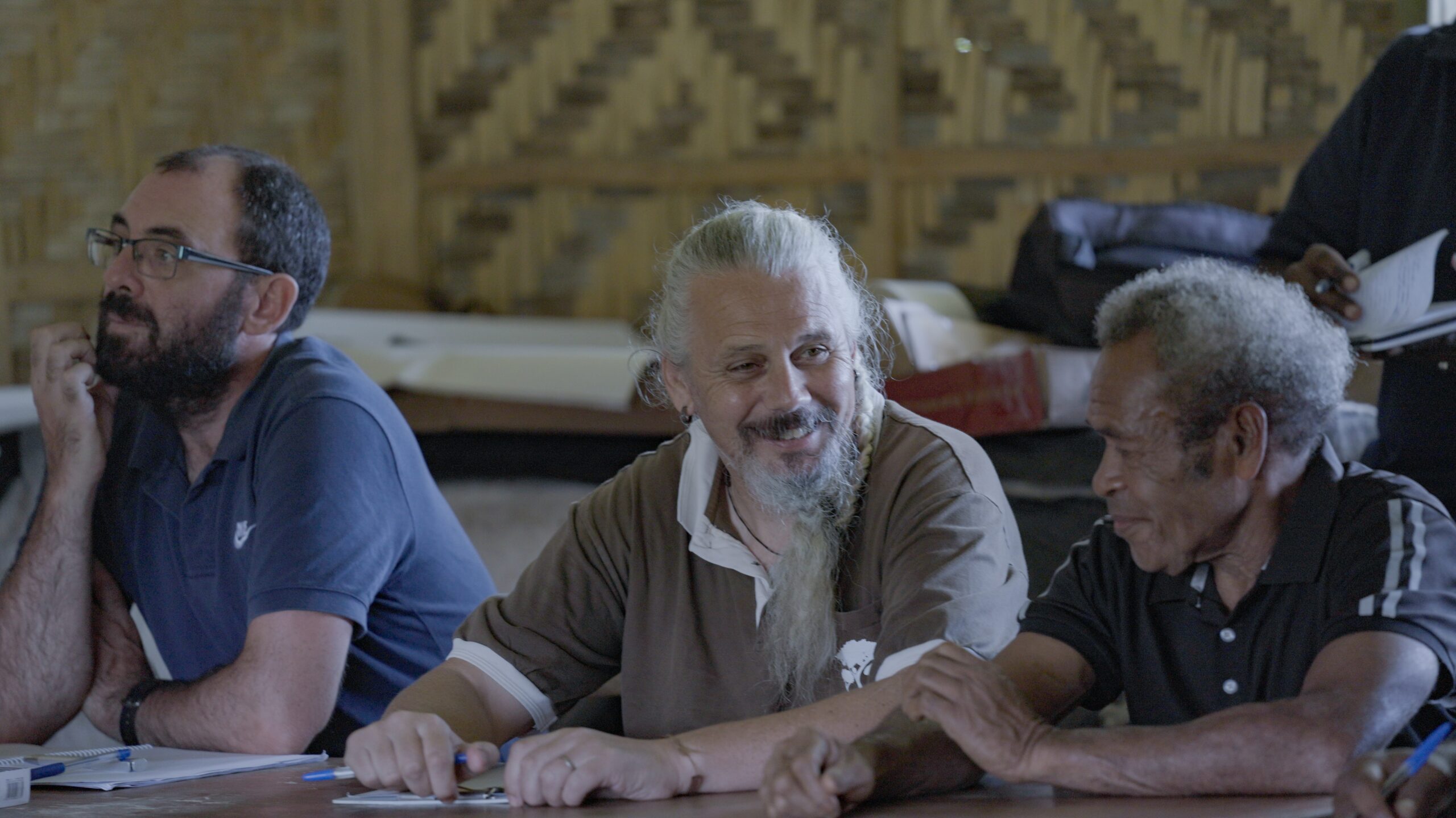 Will Unsworth, Rhett Harrison, and a local community elder sit at a wooden table during an indoor workshop, smiling and conversing with meeting papers and pens laid out before them.