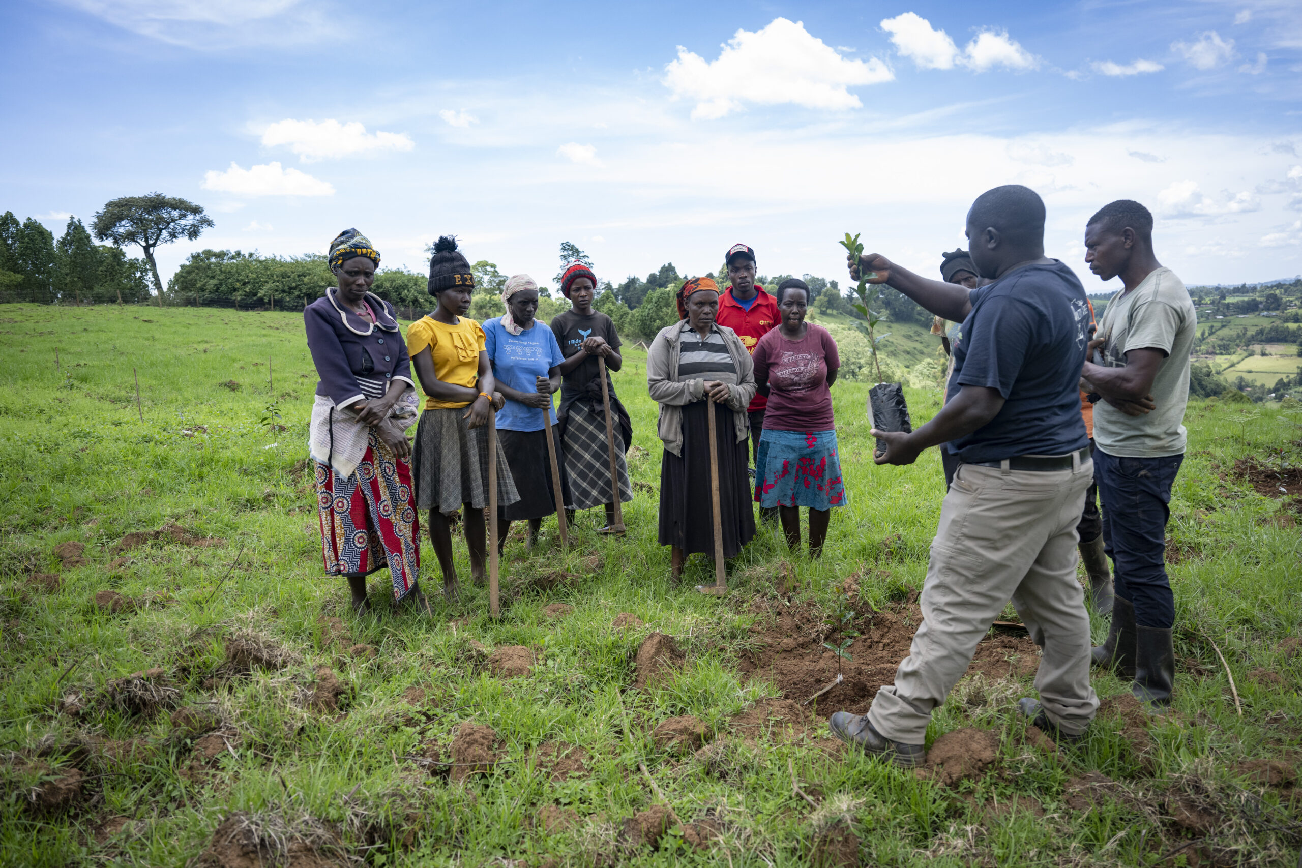 A trainer holds up a seedling while women and men watch during a field session near Maasai Mau, Kenya.