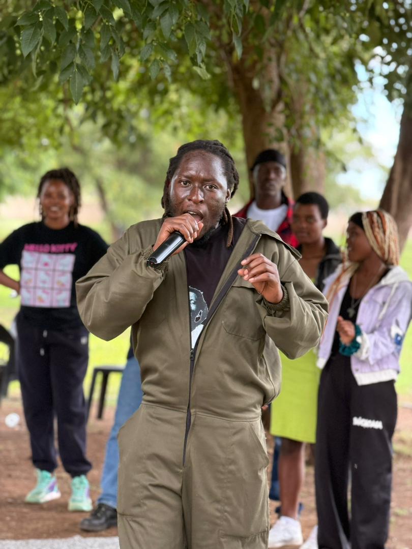 Tafadzwa Gwini, leader of Green the Ghetto, speaks into a microphone at an outdoor event, with several people standing behind him under trees.