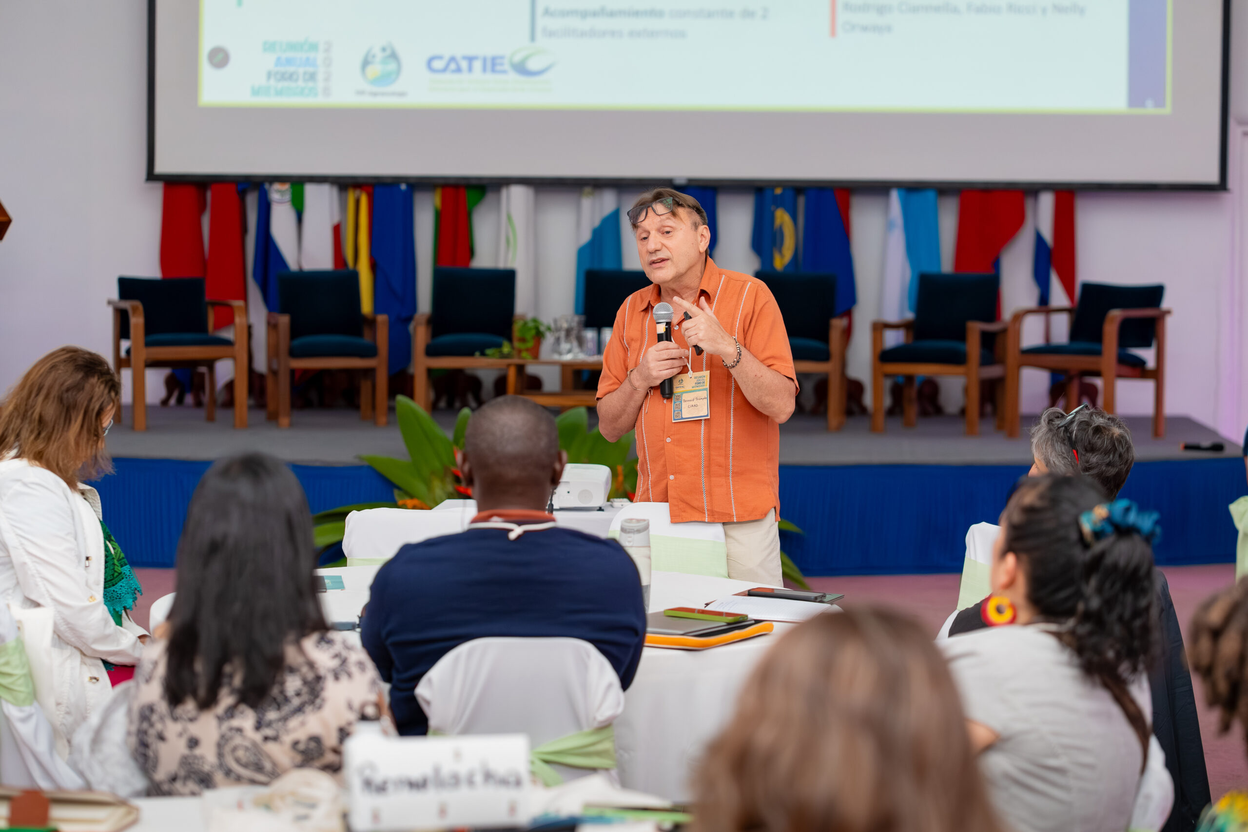 Bernard Triomphe speaks into a microphone and gestures towards participants seated around tables during the forum.