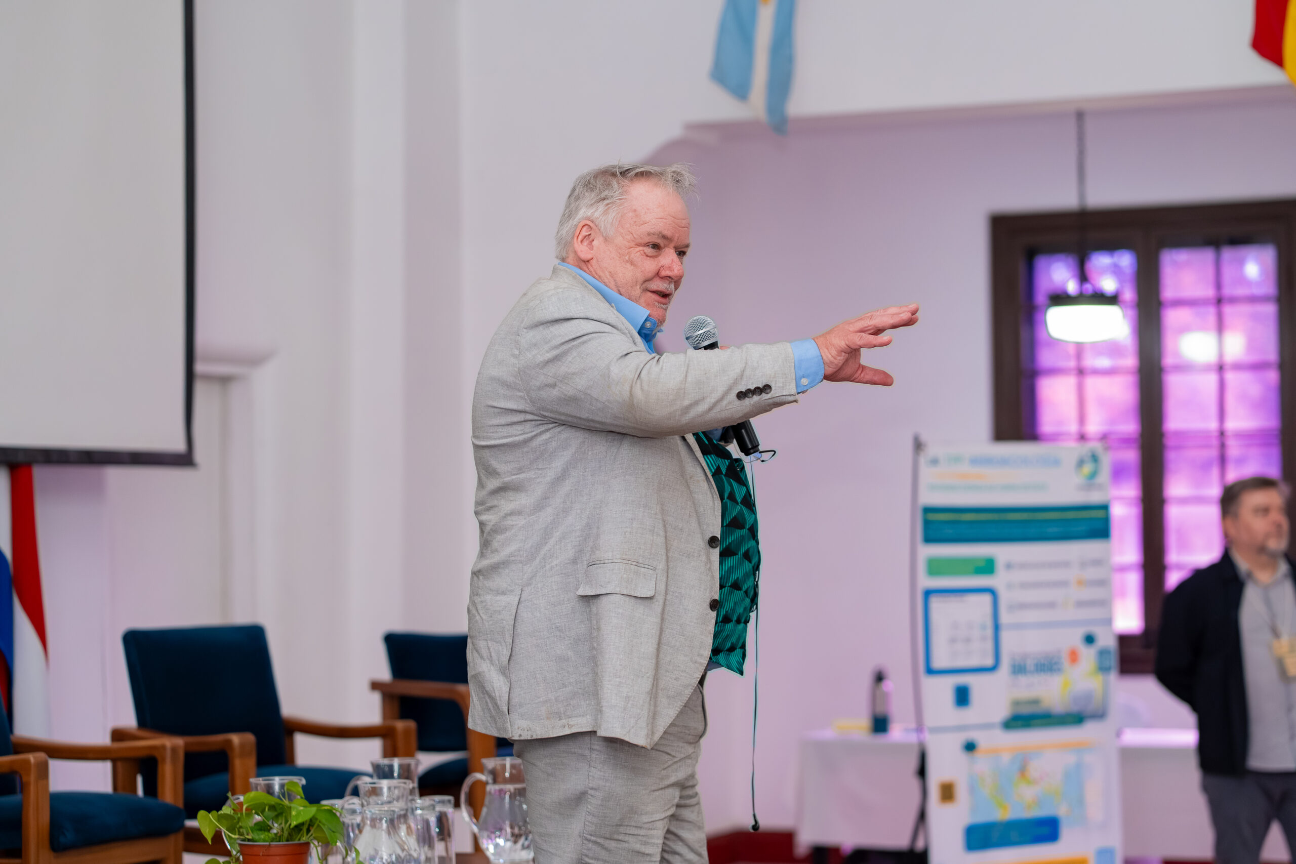 A man in a light-coloured suit speaks into a microphone and gestures towards participants during the forum.
