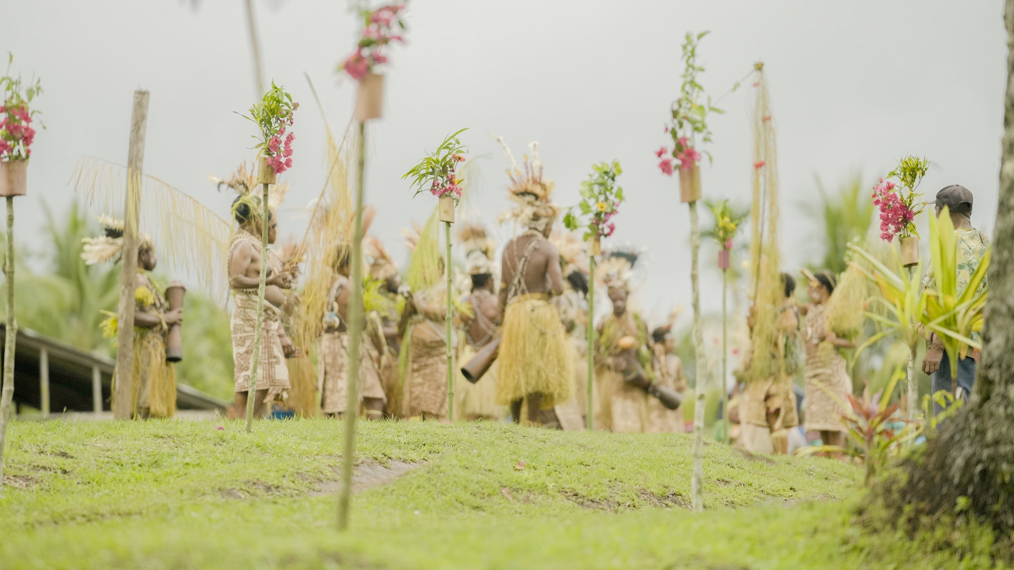 Community members in traditional dress gather on a grassy slope for a welcome ceremony in Kawowogi Village, with decorated poles and flowers in the foreground.