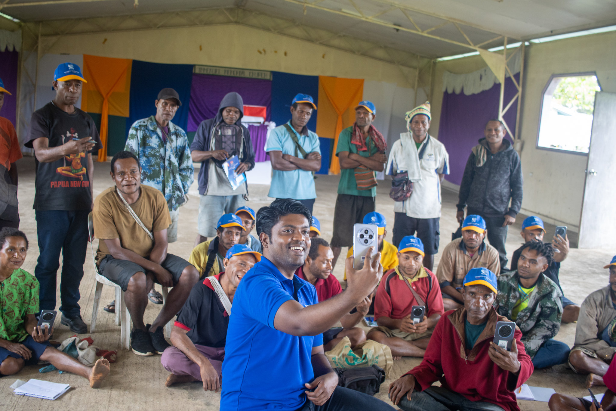 A group of participants practise taking photographs with smartphones during a social media workshop, with a facilitator demonstrating how to use a phone camera.