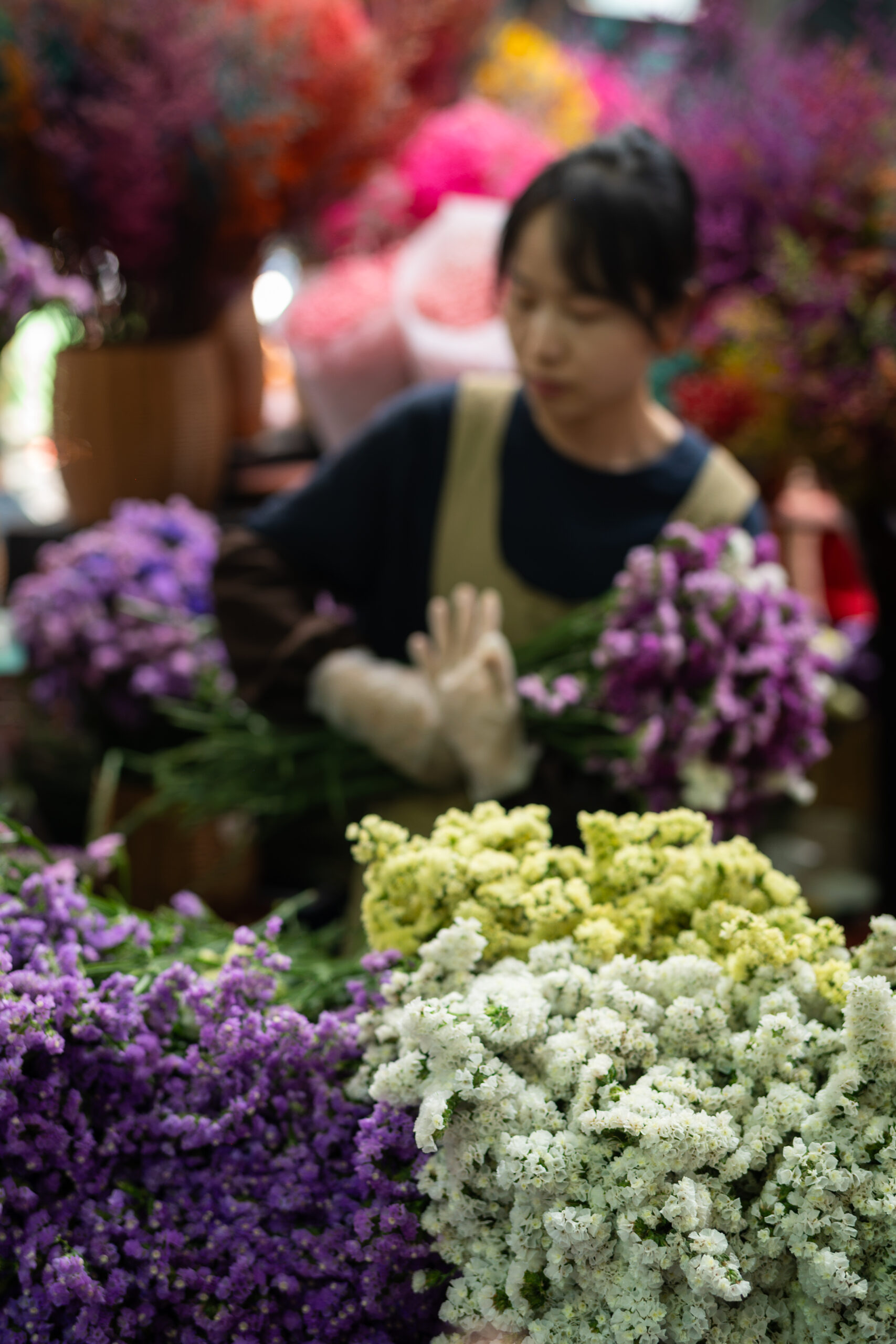A flower vendor in a dark top and apron handles bunches of purple and white flowers at Dounan Flower Market, with colourful blooms filling the background.