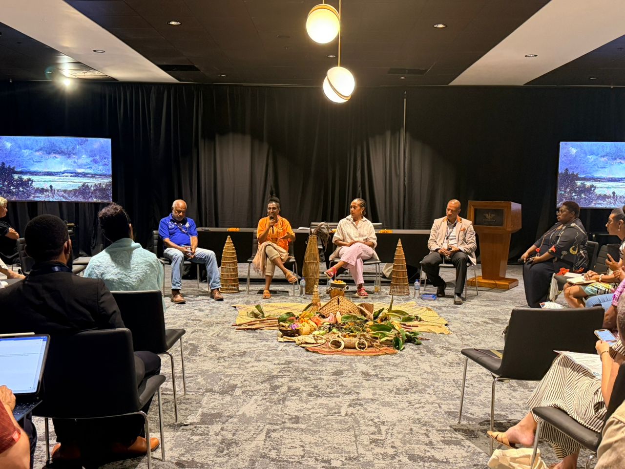Panelists speak during a session at the 2026 Melanesian Oceans Summit, with an audience seated around a display of traditional materials and a sirawa, a Binandere fishing trap, in the foreground.