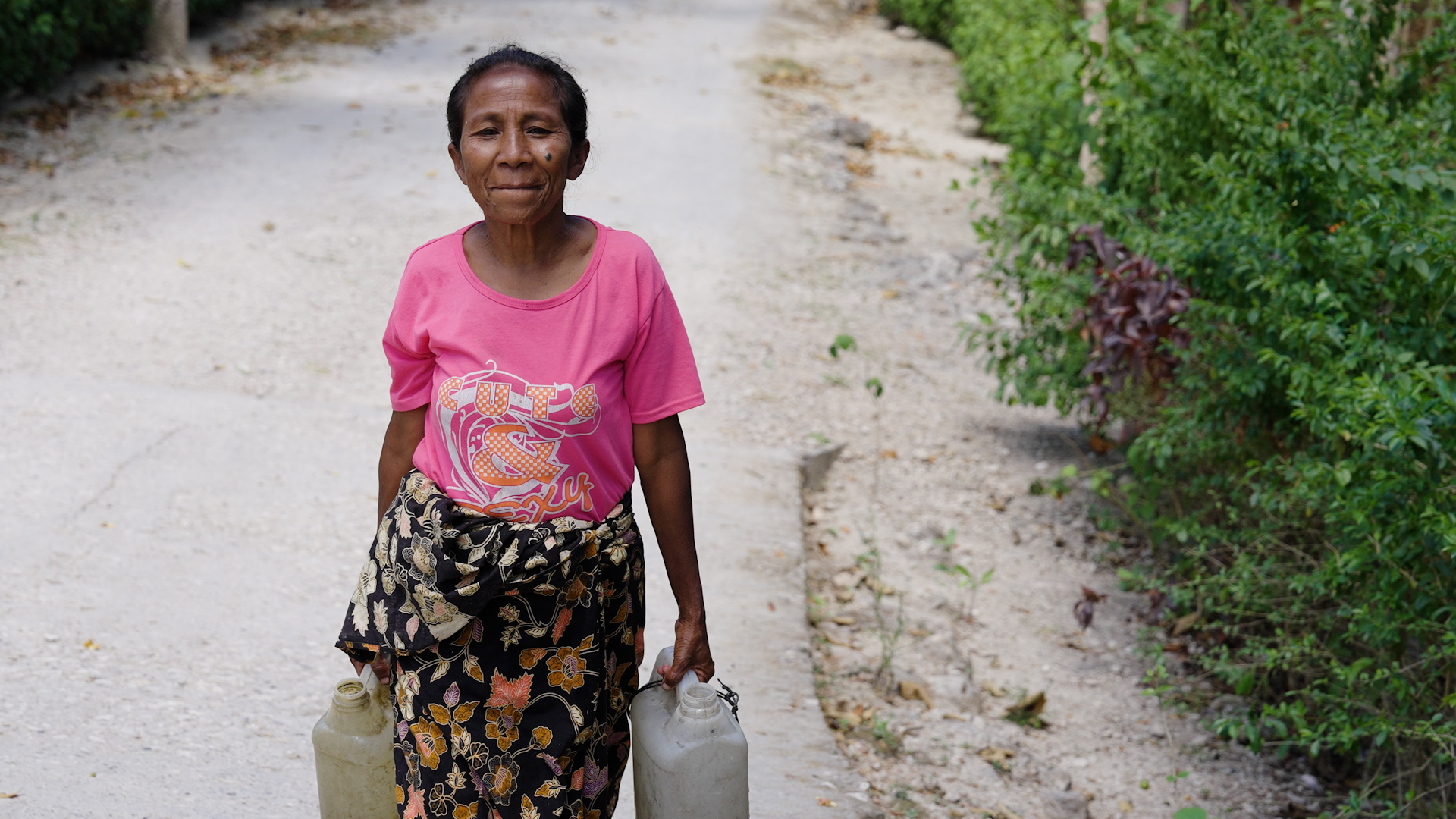 A woman in a pink shirt carries two plastic water containers along a dirt road bordered by vegetation in rural Timor, Indonesia.