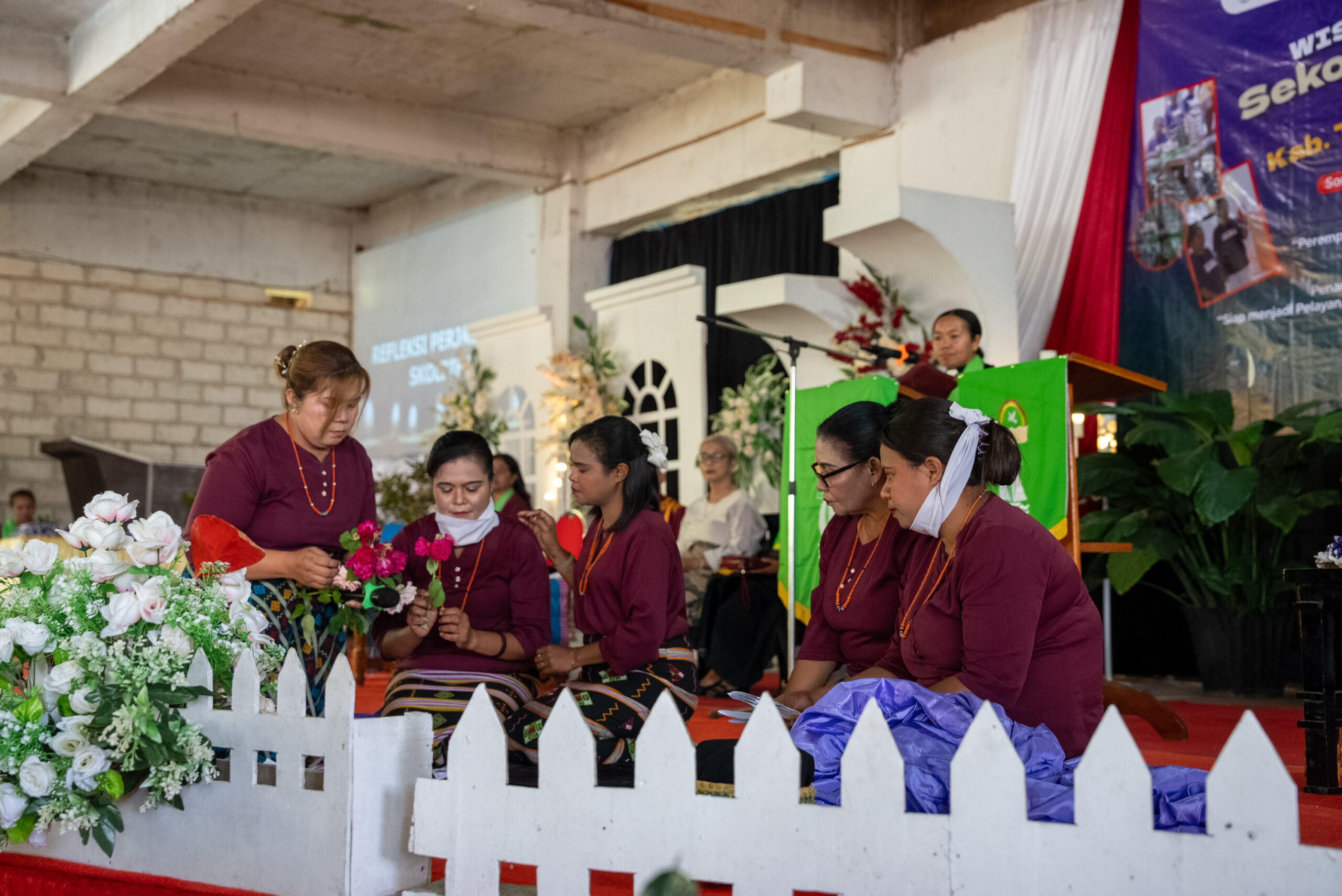 Women in traditional dress perform a theatrical scene on stage during the Skol Bife graduation ceremony, with one woman holding flowers as others sit beside her.