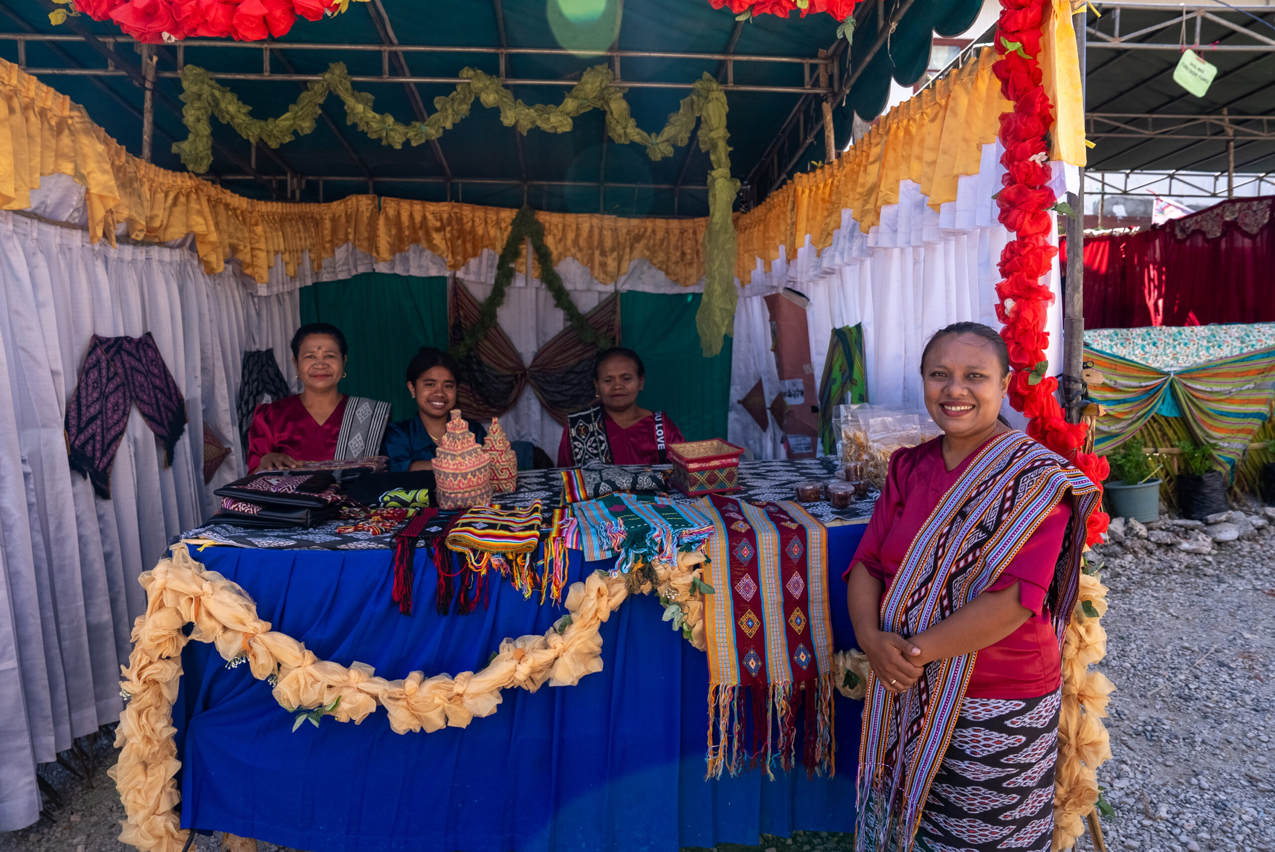 Four women stand at a Skol Bife exhibition booth displaying woven textiles, traditional handicrafts and packaged products.