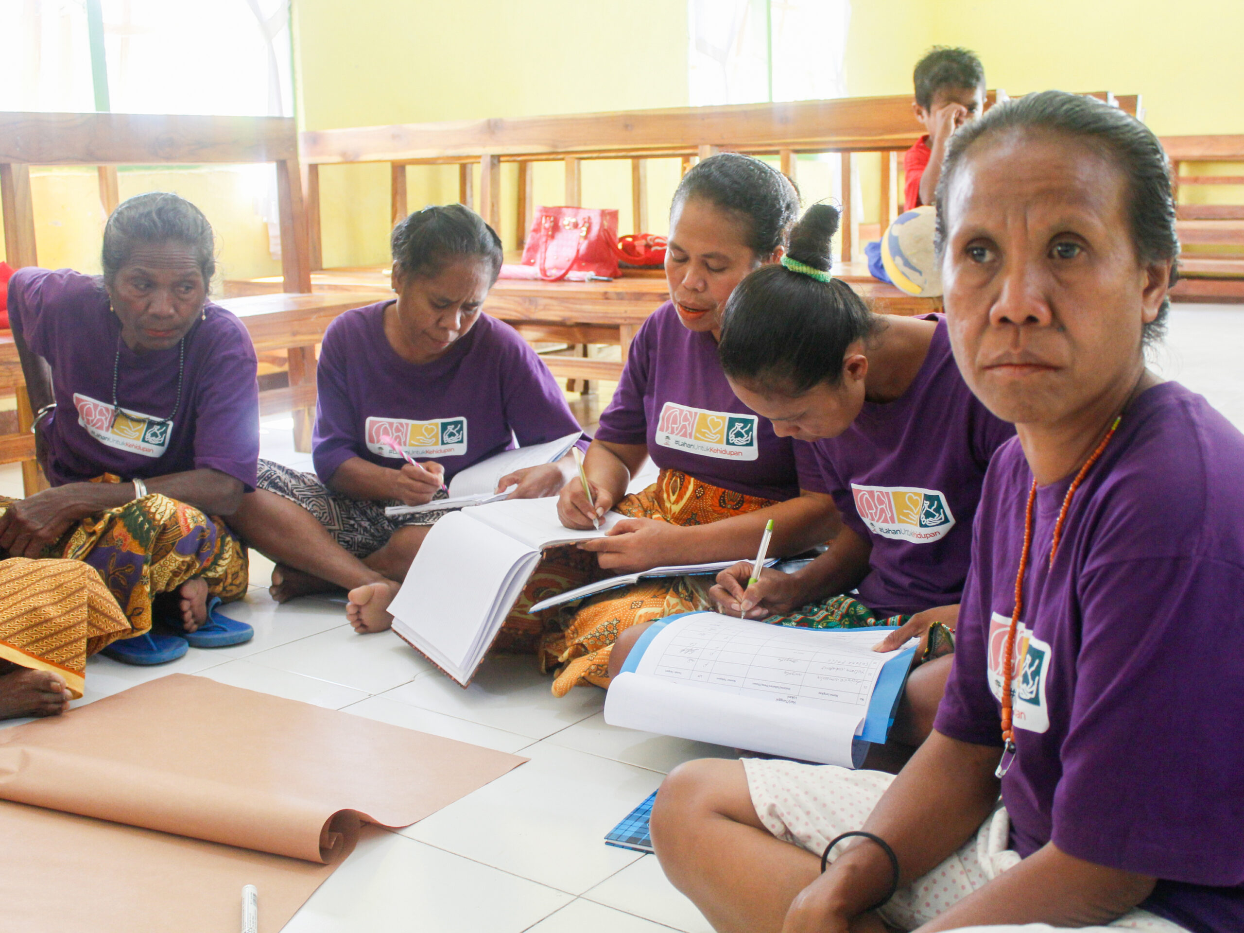 Women participating in a Skol Bife field school sit together on the floor, writing in workbooks and discussing exercises during a training session in Timor, Indonesia.
