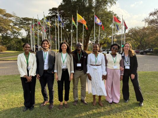 Eight panelists from the SBSTTA28 side event on sustainable wildlife management pose for a group photo outdoors, standing in front of flags representing different countries and organizations.
