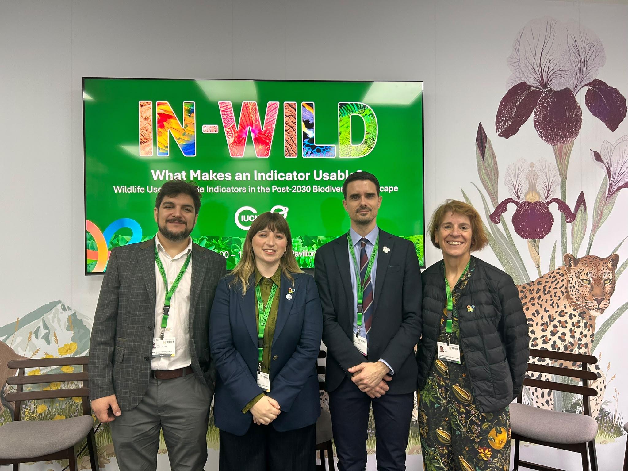 Four people stand in front of a screen displaying the IN-WILD project presentation at COP17, with botanical and wildlife illustrations on the wall behind them.