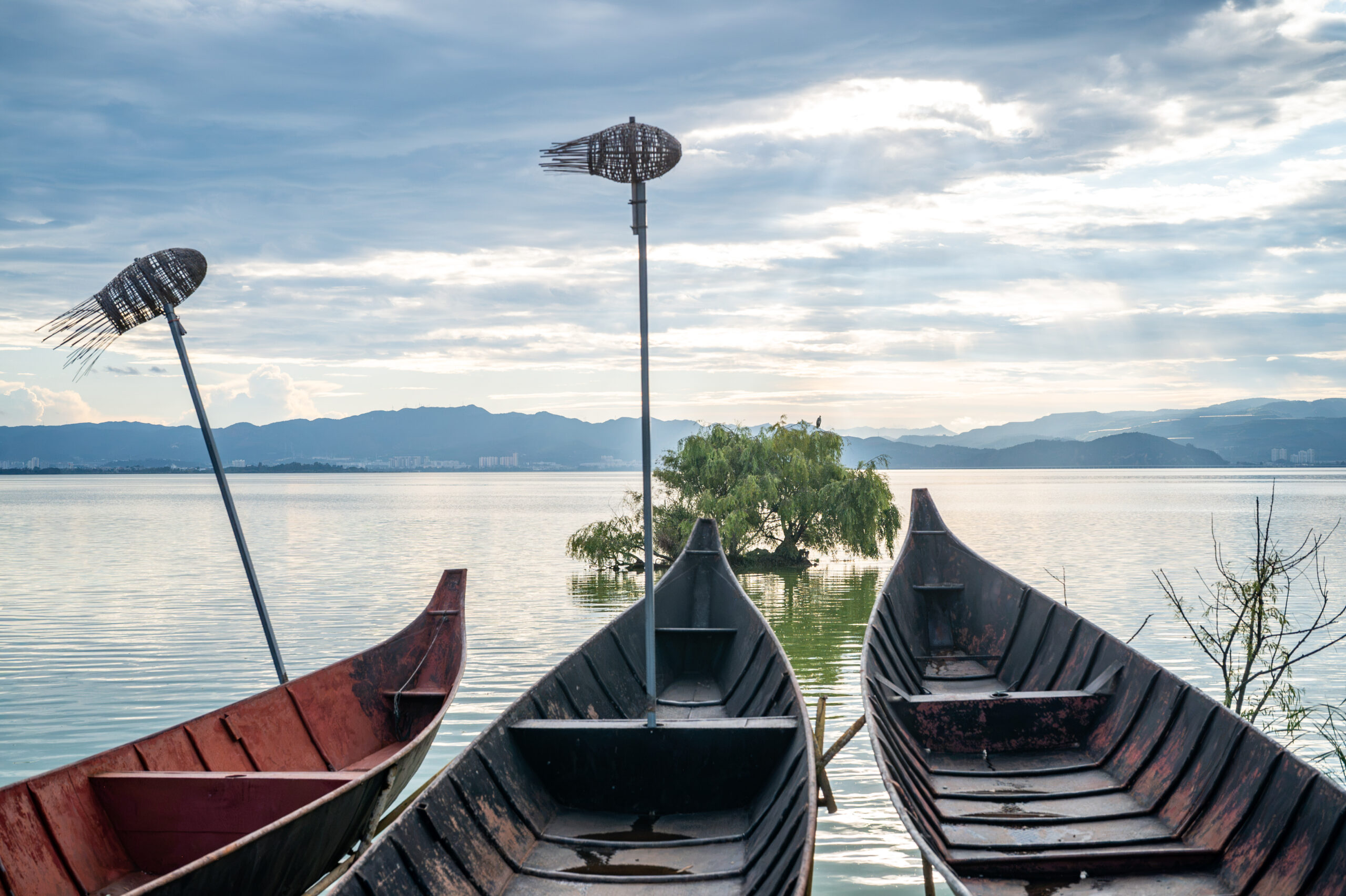 Three wooden boats rest in the calm waters of Dianchi Lake, with a small tree growing in the shallows, mountains on the horizon and a cloudy sky overhead.