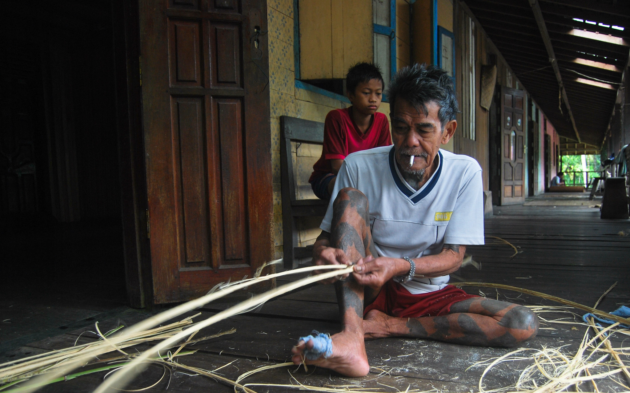 Man weaving or cleaning bemban reeds inside a traditional longhouse corridor in Kalimantan, Indonesia.