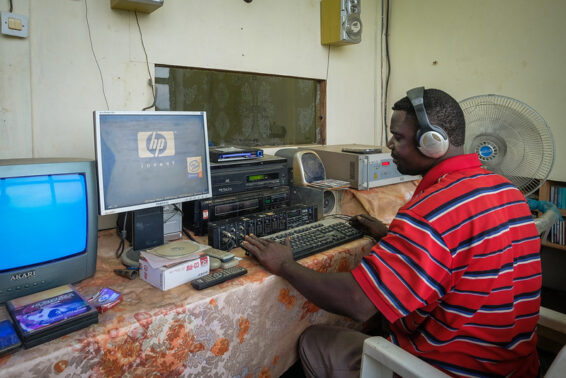 A man wearing headphones works at a radio studio desk with audio equipment and a computer in Yangambi, Democratic Republic of Congo.