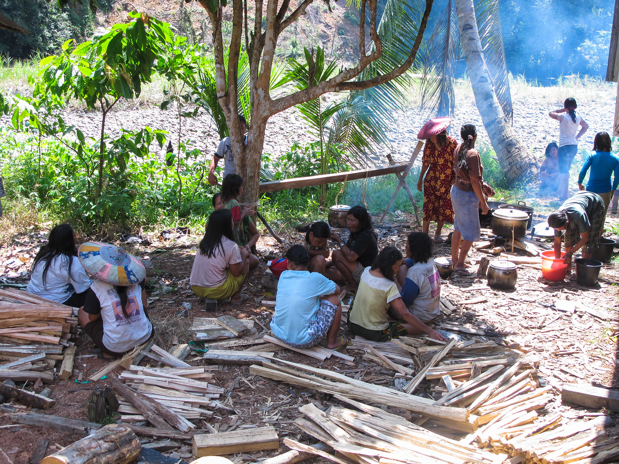 Group of villagers cooking outdoors under trees in East Kalimantan, Indonesia, with firewood and cooking pots.