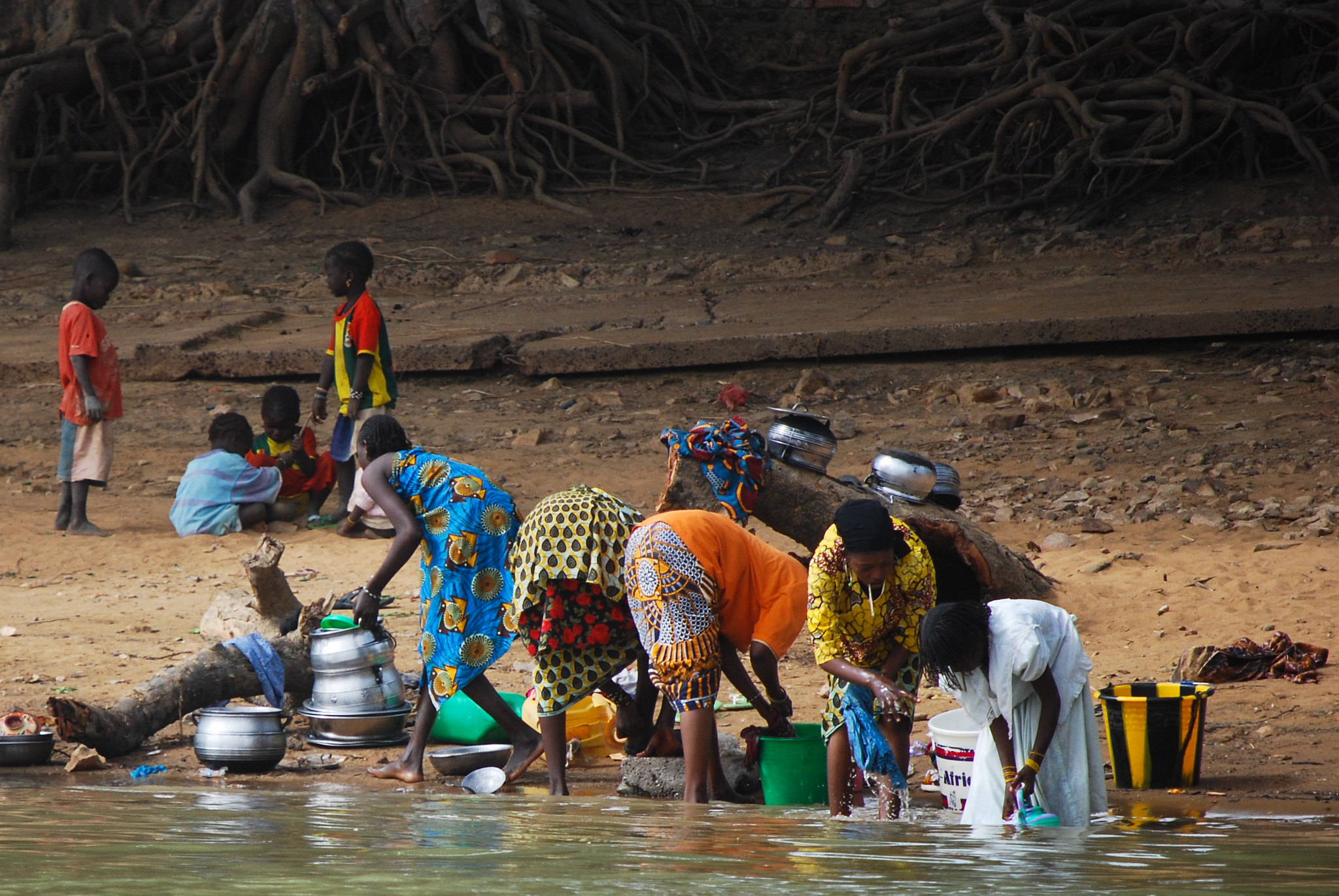 A group of women wash dishes on a riverbank in West Africa as children play nearby, reflecting the role of local communities and livelihoods in shaping equitable climate action.