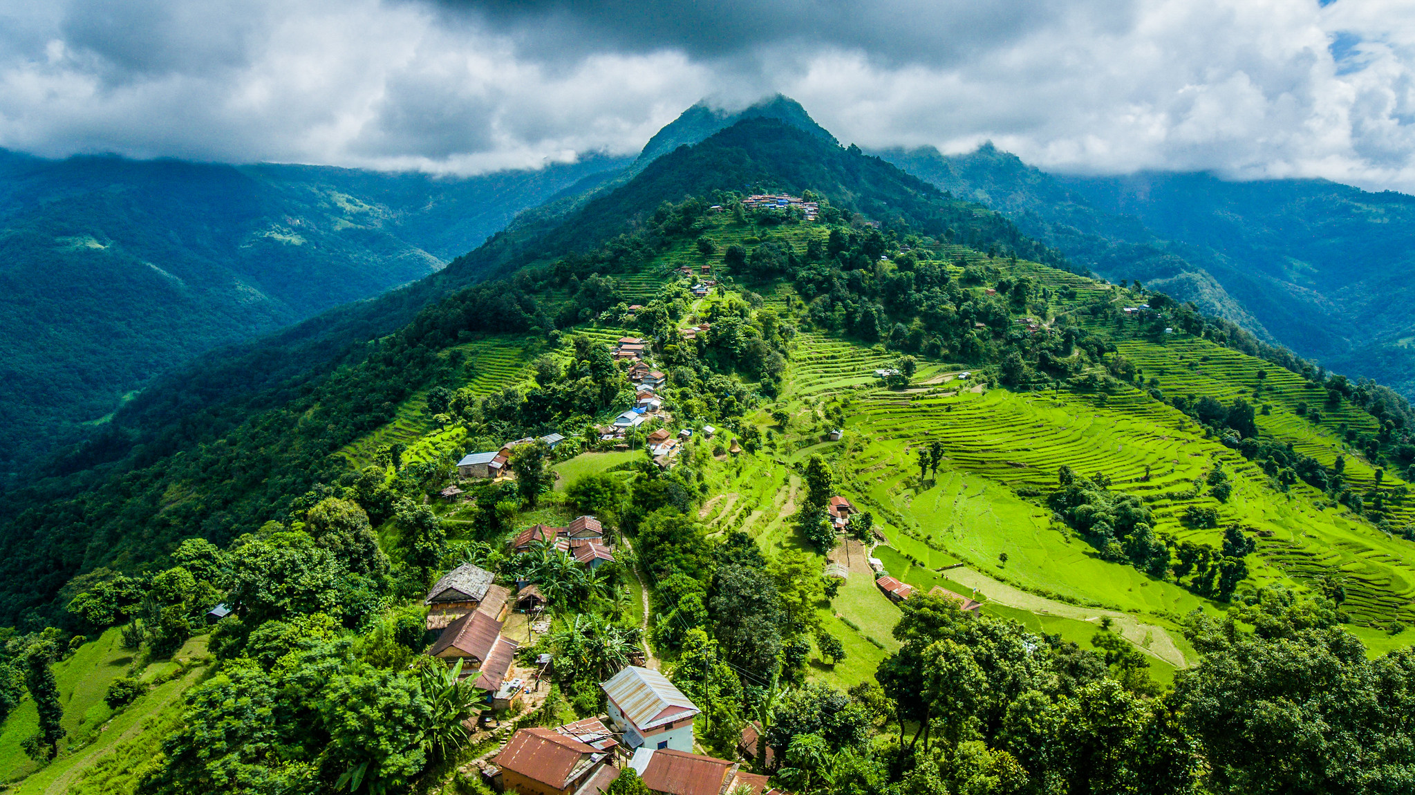Aerial view of terraced farmland, trees and village houses across steep hillsides in Nepal.