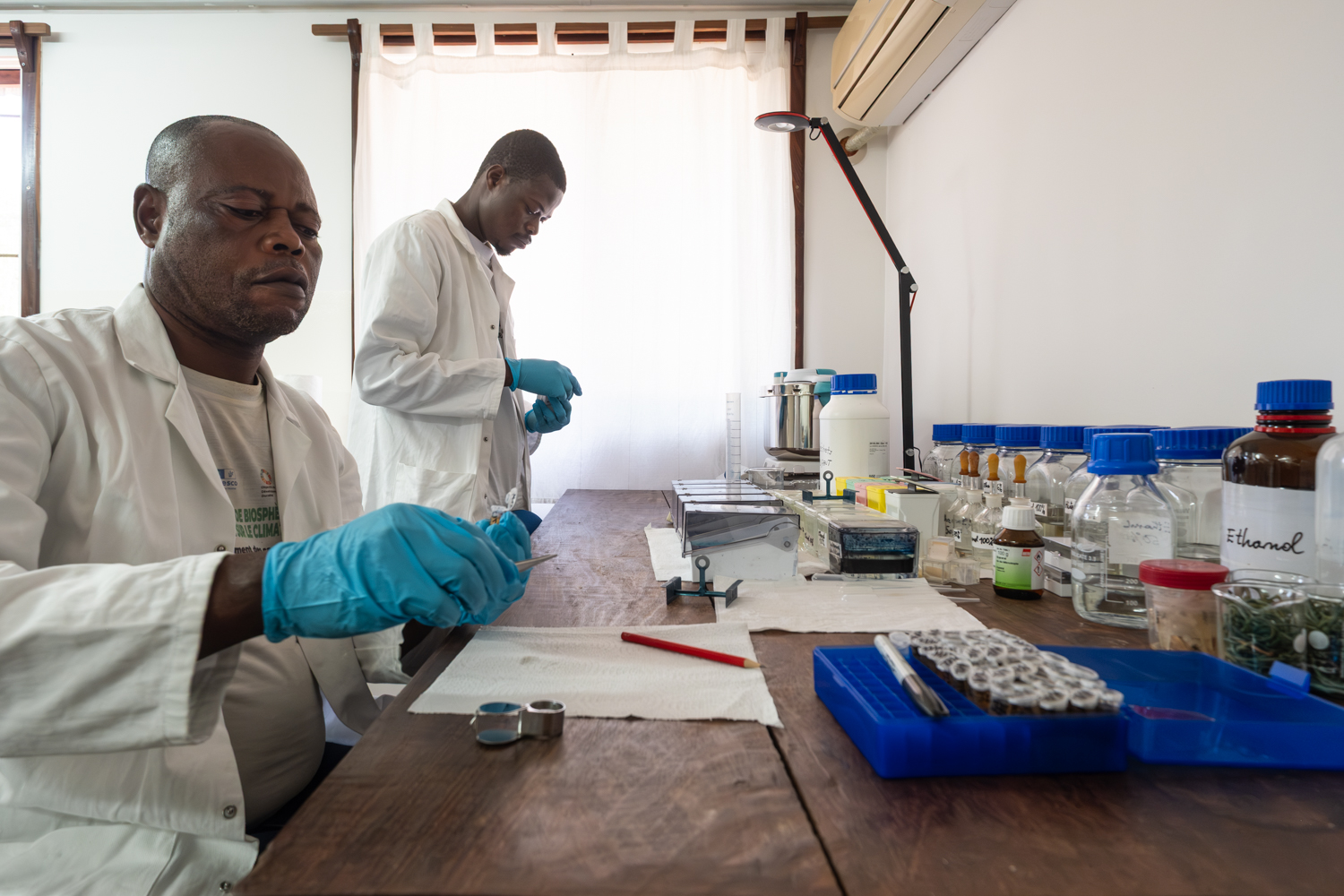Two researchers in lab coats preparing samples at a workbench with bottles and equipment.